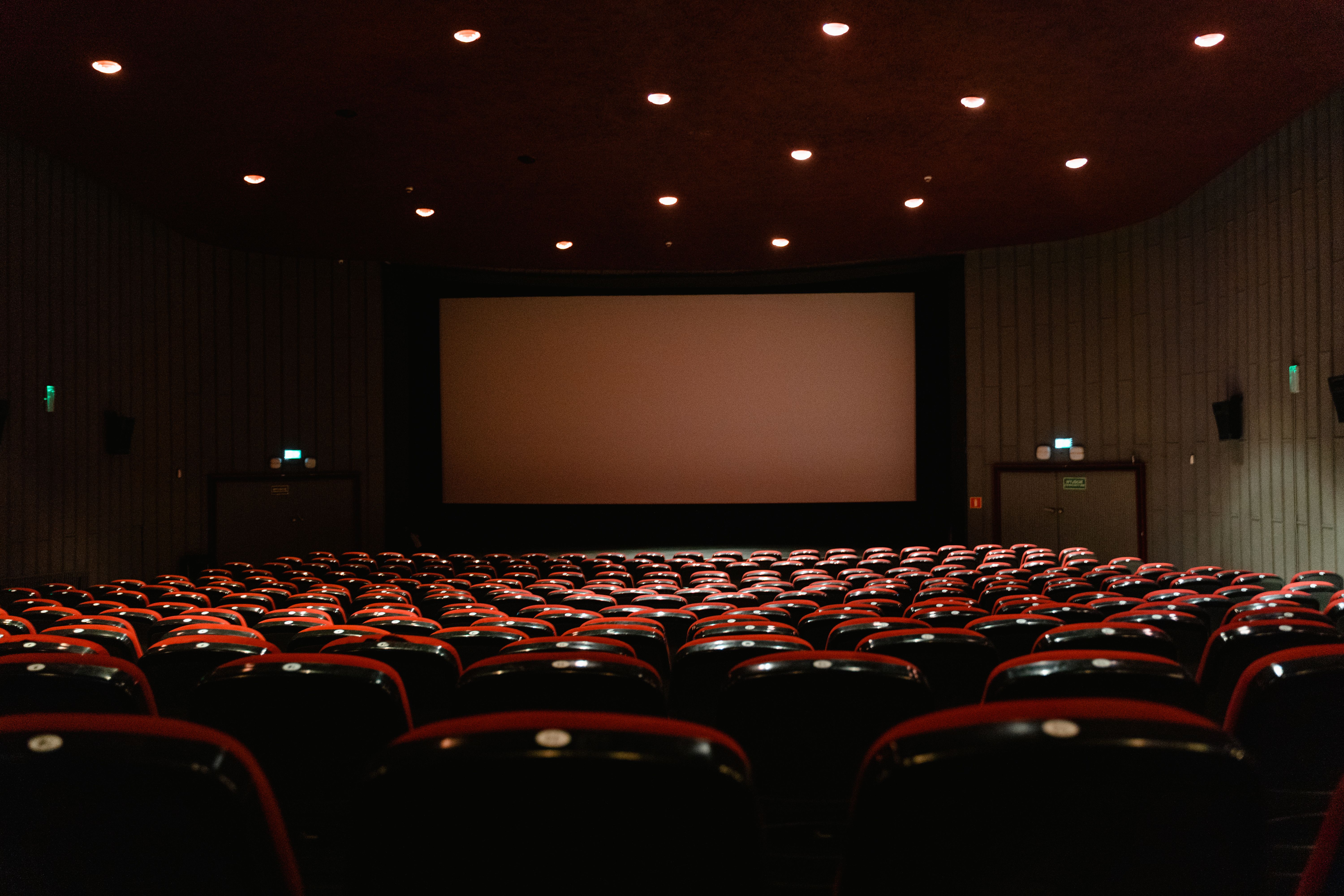 A dimly lit empty cinema theater with rows of red chairs facing a projector screen, evoking a night at the movies