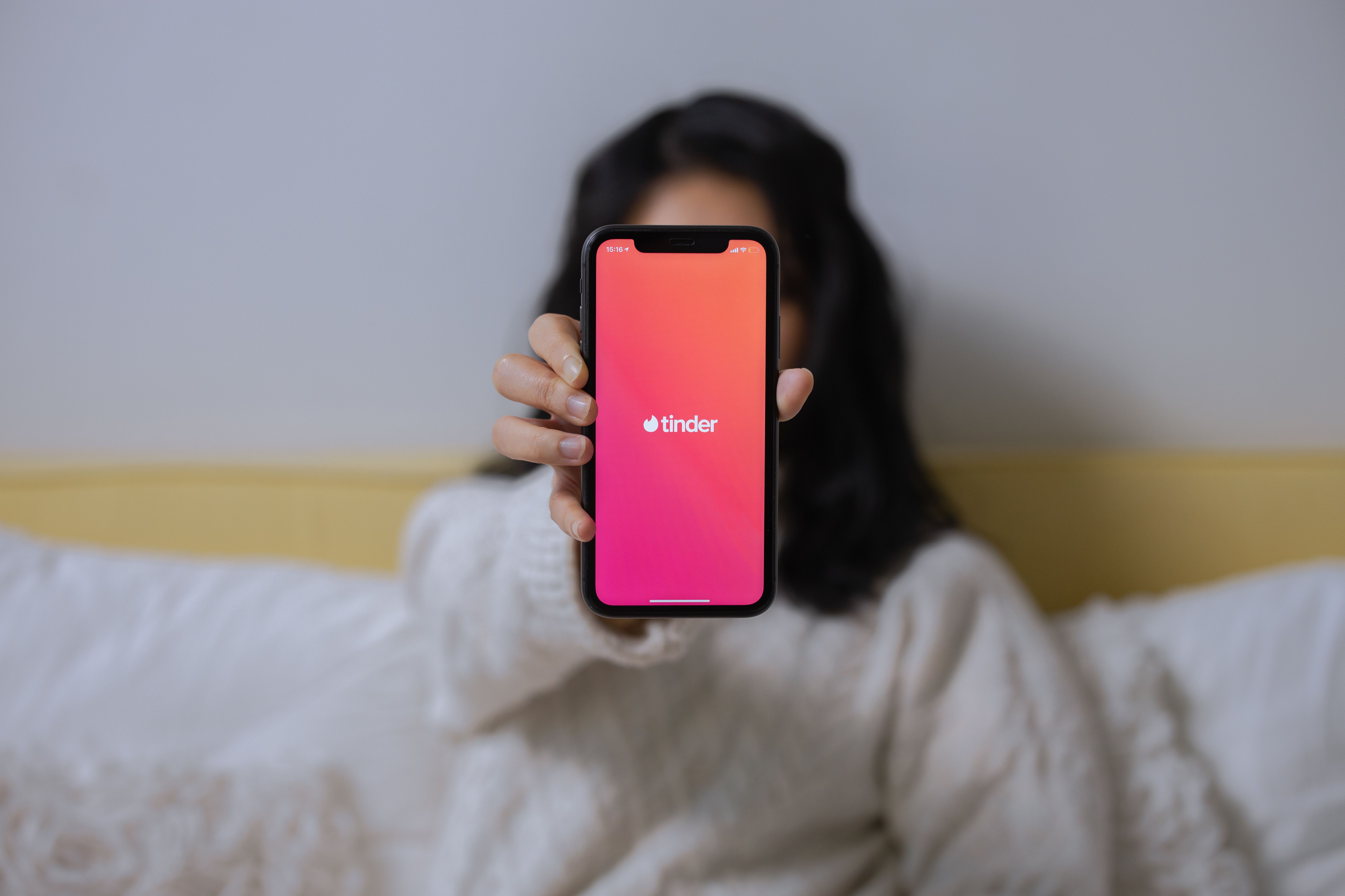 A woman displays a dating app on her smartphone while sitting indoors, illustrating app-based and commodified romance.