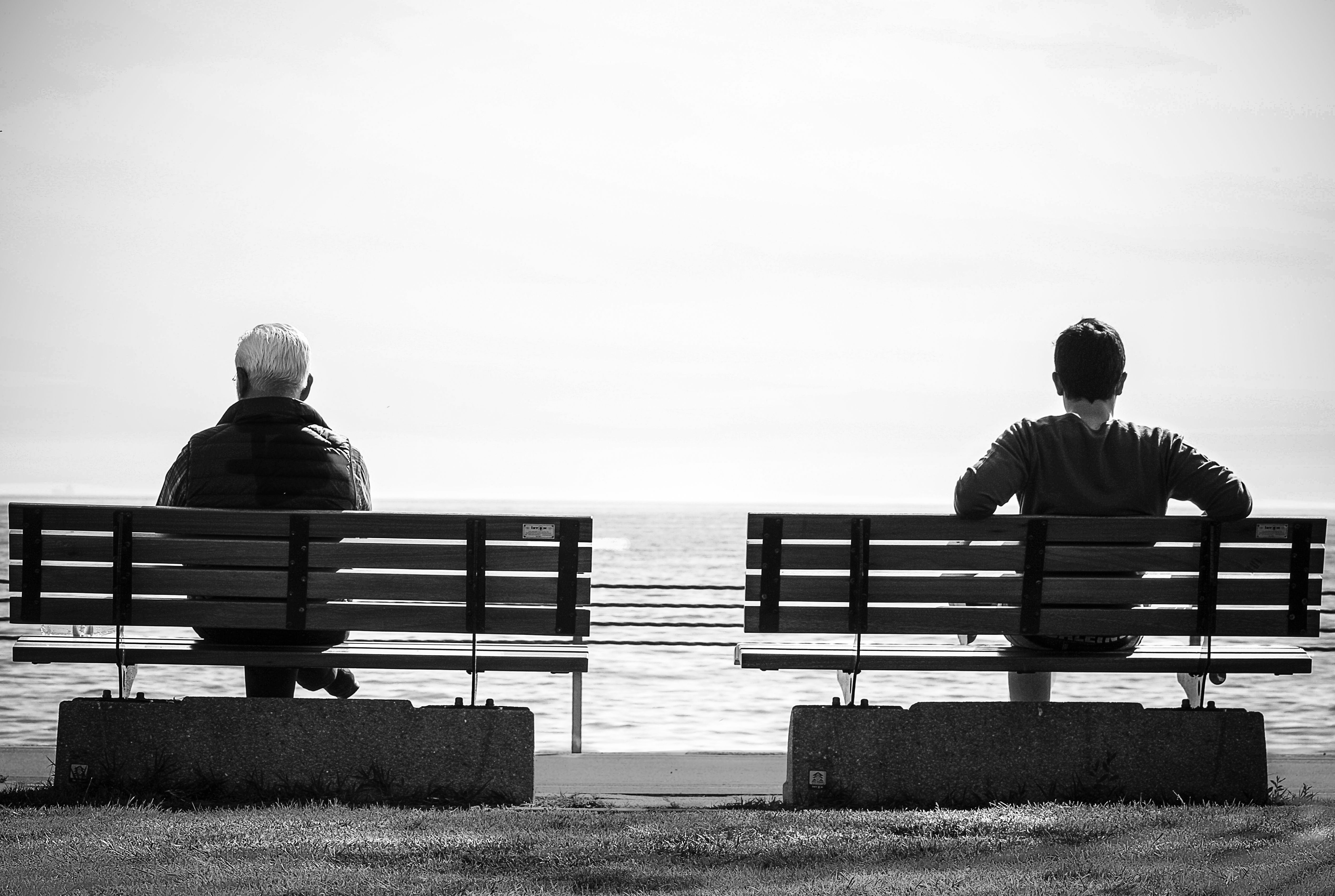 Black and white photo of two people sitting on separate benches by the sea, symbolizing emotional distance.