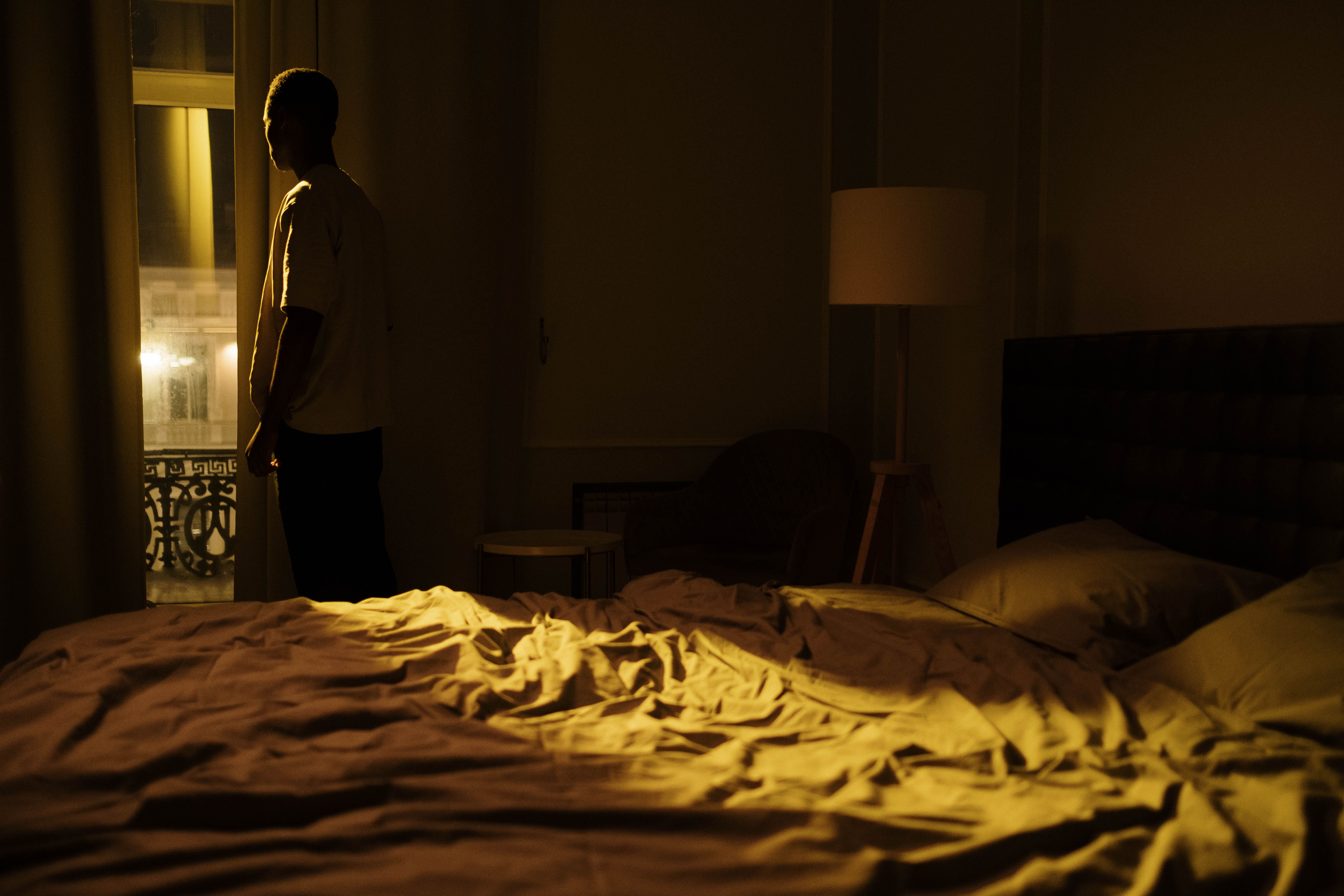 A man sits in a dark bedroom gazing out the window at night, capturing male solitude and loneliness.