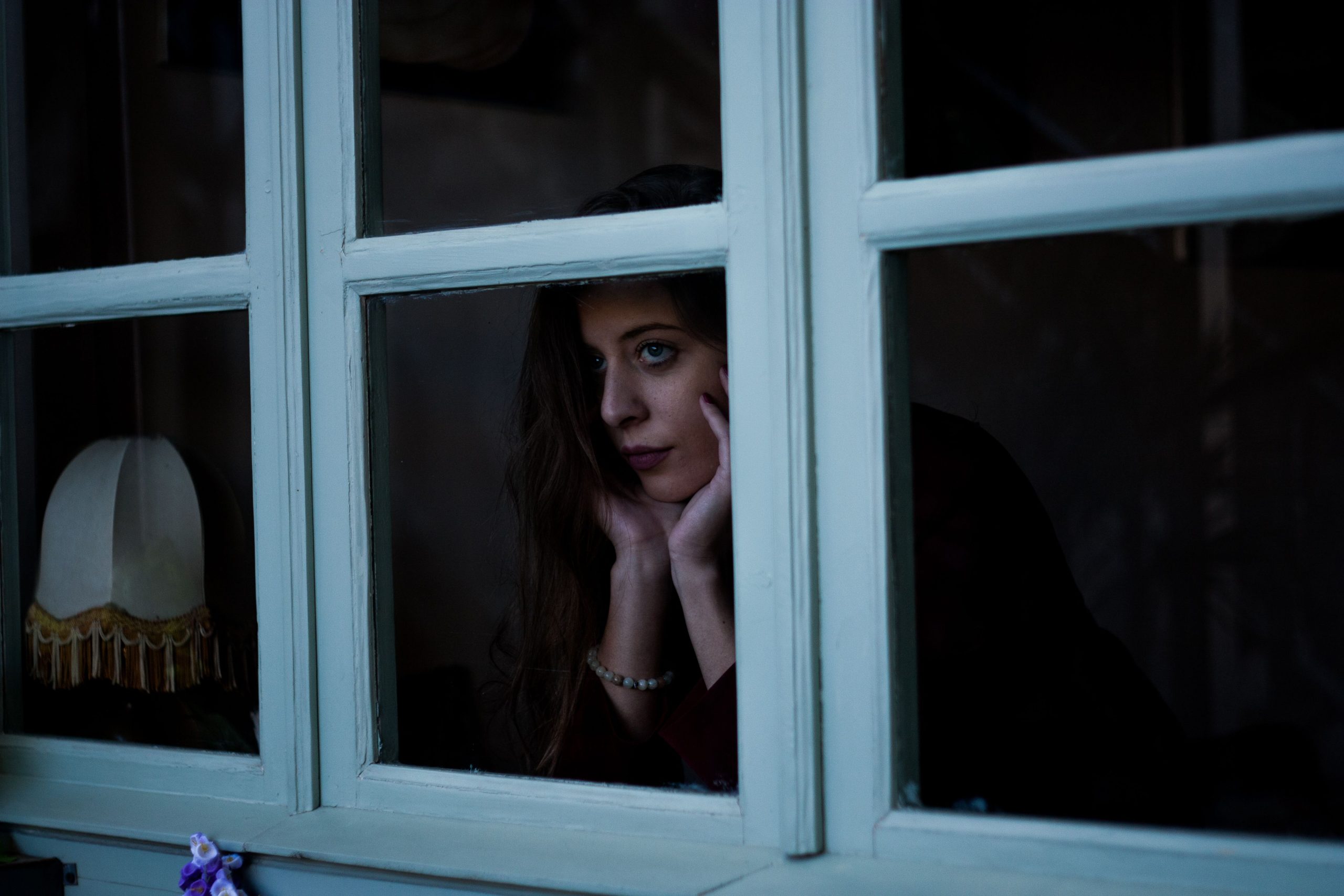 A woman gazes thoughtfully out a window at night, a moody and introspective scene that evokes modern loneliness.