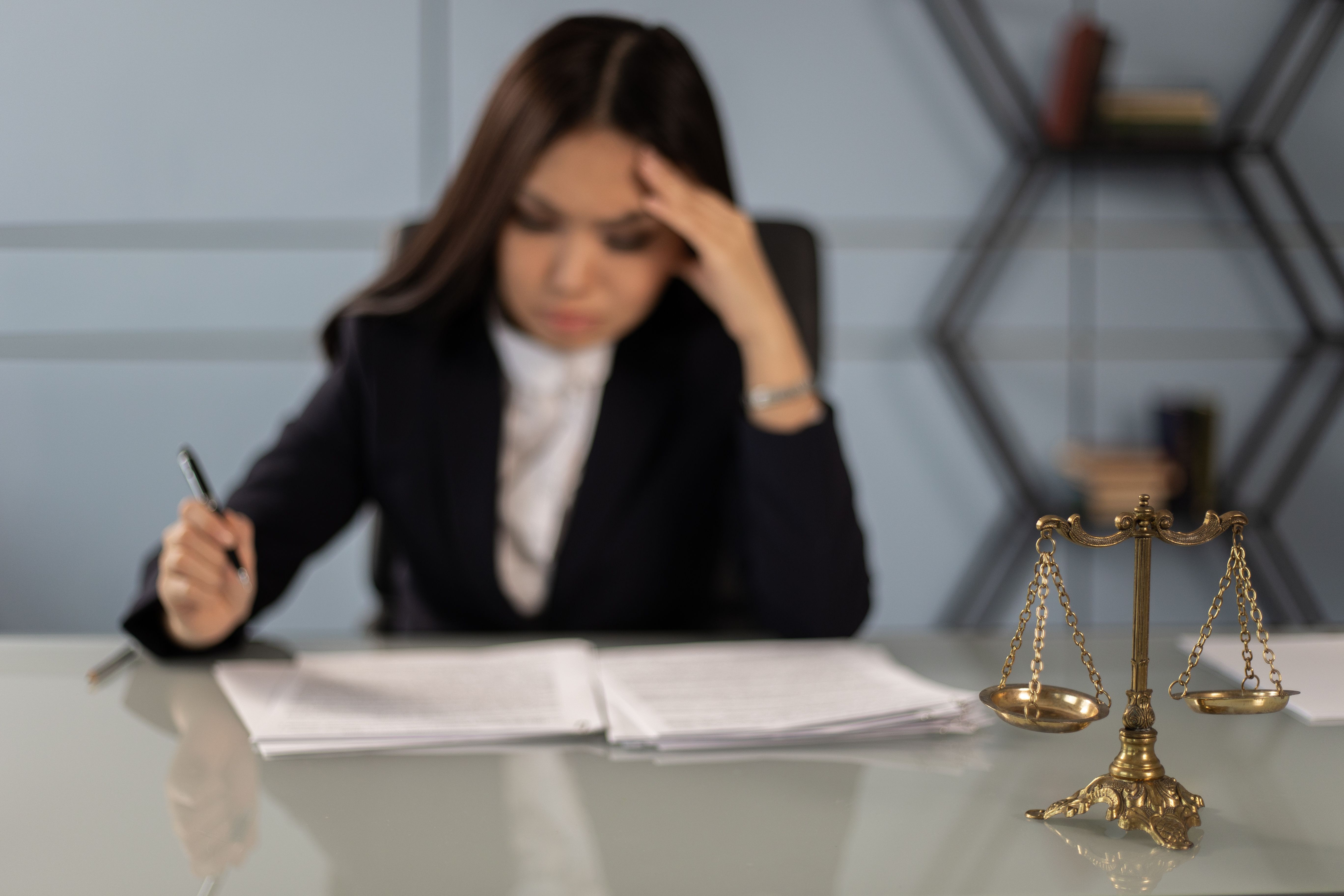 Fatigued female lawyer reviewing documents beside a justice scale, capturing stress in a crumbling legal workplace